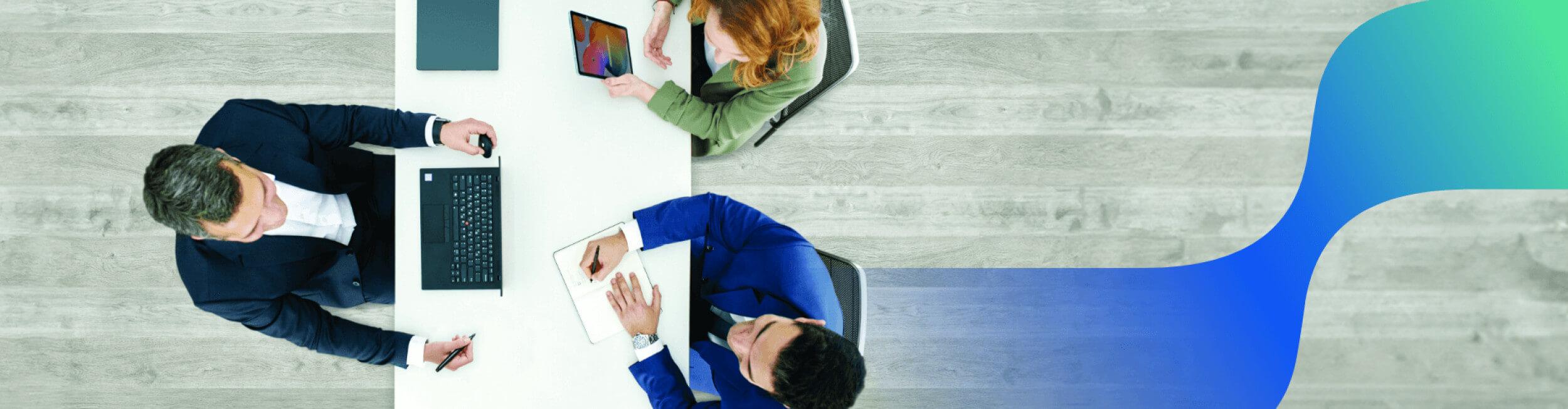 τρεις άνθρωποι κάθονται σε γραφείο με laptop, tablet και σημειωματάριο και συζητάνε